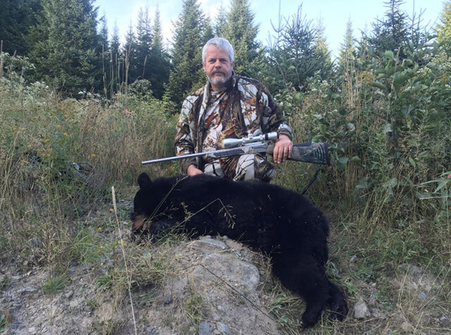 far away shot of man posing with dead black bear on stump